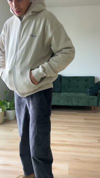 Sudadera beige con cremallera y bolsillos laterales, combinada con pantalones cargo de pana oscuros que tienen bolsillos laterales. Entorno interior con un sofá verde.