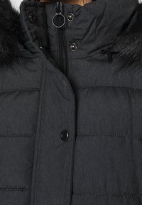 Gros plan d'une veste matelassée d'hiver foncée avec fermeture éclair, boutons-pression et capuche bordée de fourrure visible sur les bords supérieurs.