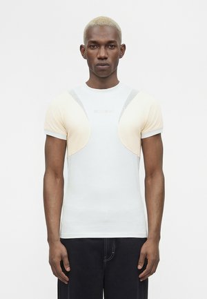 Jeune homme aux cheveux blonds courts, portant un haut blanc et beige ajusté à manches courtes et un pantalon foncé, debout devant un fond uni.