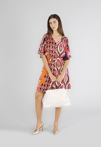 Värikäs polvipituinen mekko, jossa on V-aukko, leveät hihat ja kaleidoskooppinen kuosi. Lisävarusteina valkoinen käsilaukku ja kultaiset korkokengät.