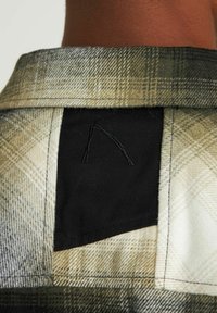 Camicia a quadri in tonalità beige, verde e nero con una toppa nera sul collo posteriore che mostra un logo stilizzato con cuciture nere.