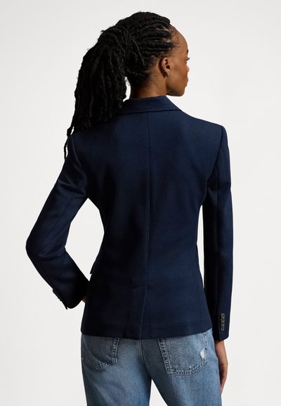Ναυτικό μπλε σακάκι με δομημένους ώμους και μονό σκίσιμο στο πίσω μέρος· διαθέτει εφαρμοστή γραμμή και μανσέτες με κουμπιά. Φορεμένο πάνω από ανοιχτόχρωμο τζιν.