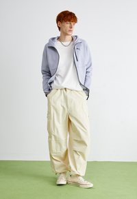 Felpa zip-up lilla con dettagli neri, t-shirt bianca, pantaloni cargo beige e sneakers beige con strisce multicolori.
