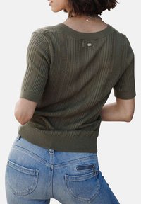 Pull en tricot vert olive à manches courtes, texture côtelée, et détail de bouton au dos, associé à un jean bleu clair.