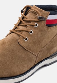 Brauner Wildleder-Stiefelett mit hellbraunen Schnürsenkeln, Metallösen, gestreiftem Stoffkragen und weißer sowie schwarzer Gummisohle.