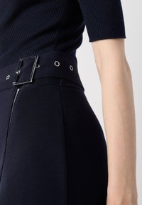 Pantalons taille haute bleu marine avec une large ceinture, dotée d'une boucle en argent brillant et d'œillets, tissu texturé et fermeture éclair latérale.