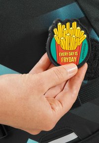 Gummi-Patch mit einem roten Behälter für Pommes Frites mit gelben Akzenten und dem Text "JEDER TAG IST POMMESTAG", gehalten gegen eine strukturierte schwarze Oberfläche.