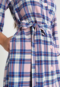 Rosa karierter Kleid mit blauen, gelben und roten Mustern. Es hat einen gebundenen Gürtel, Knöpfe und Seitentaschen. Weicher Stoff mit lockerer Passform.