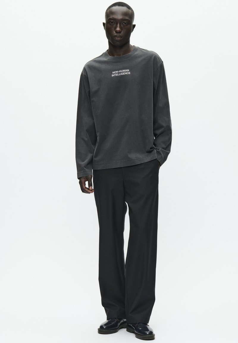 Chemise en coton à manches longues, gris foncé, avec le texte "INTELLIGENCE NON HUMAINE", associée à un pantalon noir ample et des chaussures noires. Texture lisse.
