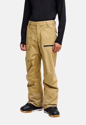 Persoon draagt losse beige cargobroek met zwart ritszakdetail en zwarte schoenen, getoond van midden torso tot voeten tegen een witte achtergrond.