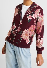 Giacca bomber bordeaux con fiori rosa e verdi, chiusura con zip, polsini e orlo a coste, realizzata in tessuto morbido e testurizzato.