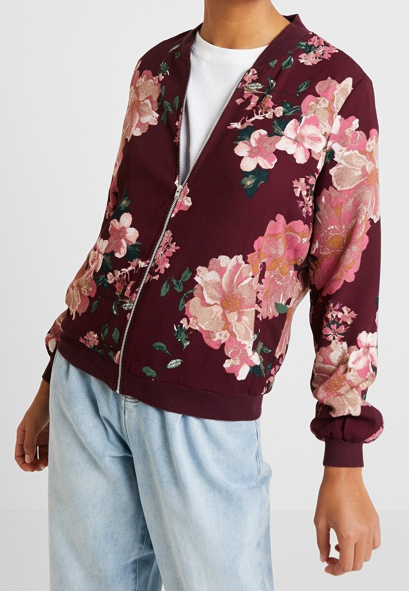 Giacca bomber bordeaux con fiori rosa e verdi, chiusura con zip, polsini e orlo a coste, realizzata in tessuto morbido e testurizzato.