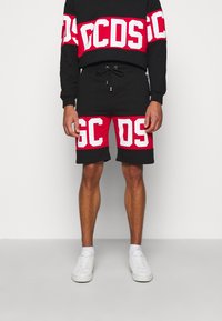 Homme portant un short sportif noir et rouge et un sweat-shirt avec de grandes lettres blanches en bloc, des baskets blanches, debout devant un fond blanc.