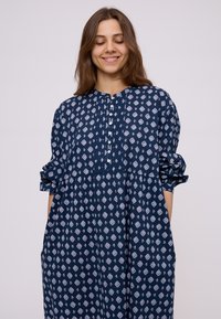 Giovane donna con capelli castano medio che indossa un vestito blu navy largo con un motivo geometrico bianco, sorridente con gli occhi chiusi.