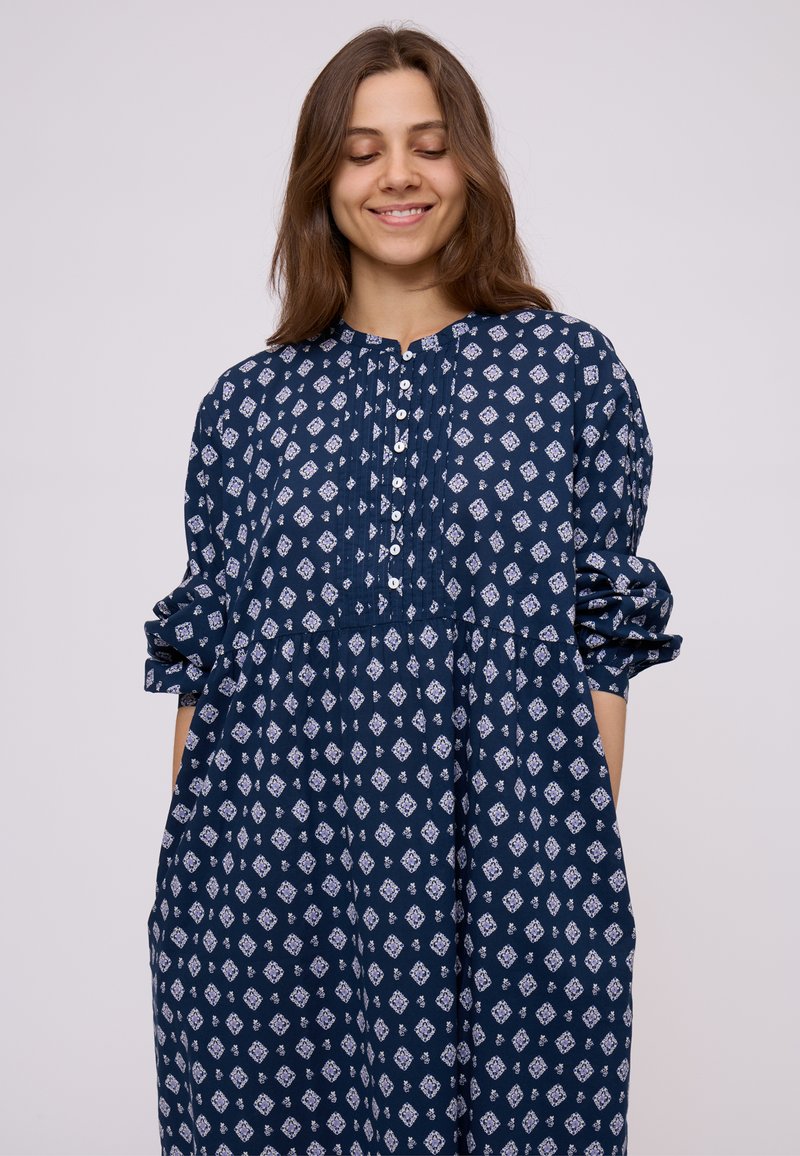 Giovane donna con capelli castano medio che indossa un vestito blu navy largo con un motivo geometrico bianco, sorridente con gli occhi chiusi.
