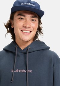 Námořnická modrá mikina s šedými šňůrkami má vyšitý logo "Quiksilver" v růžové barvě. Tkanina vypadá měkce a má hladkou strukturu.