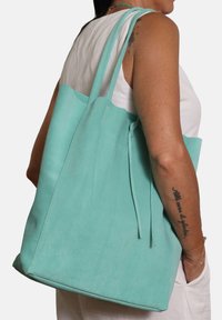 Een muntgroene suède schoudertas met twee handvaten en een striksluiting. Het oppervlak heeft een gladde textuur en een afgeronde vorm.