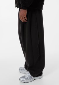 Schwarze, locker geschnittene Hose mit weitem Bein, aus weichem Stoff und mit subtilen Falten. Kombiniert mit metallisch silbernen Sportschuhen.