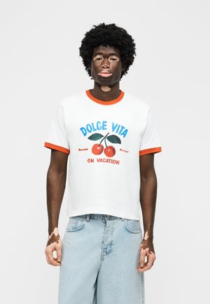 Młody mężczyzna z bielactwem w białej koszulce z pomarańczowym wykończeniem i grafiką wiśni z napisem "Dolce Vita na wakacjach", w połączeniu z jasnoniebieskimi dżinsami.