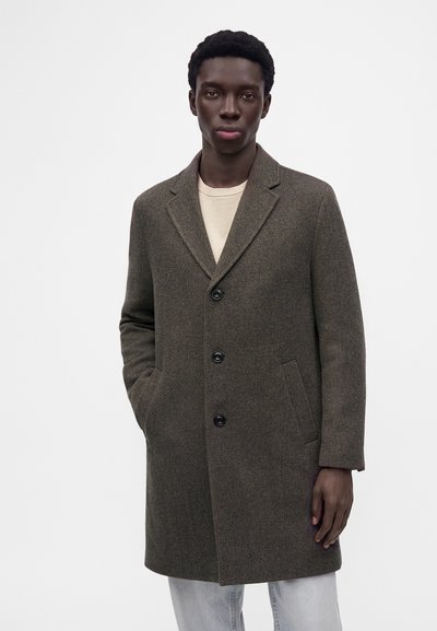 Homme portant un pardessus marron à trois boutons sur une chemise beige, les mains dans les poches du pardessus, debout devant un fond blanc uni.