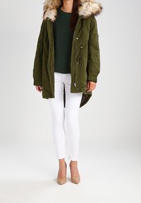 Olivgrüne Parka-Jacke mit Kunstpelz-Kapuze, Frontreißverschluss und Kordelzug, kombiniert mit weißen Skinny-Hosen und beigen High Heels.
