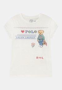 Biała dziecięca koszulka z krótkim rękawem z haftowanym tekstem "Polo Ralph Lauren", małą kwiatką oraz niedźwiedziem w zielonym swetrze, dżinsach i tenisówkach.