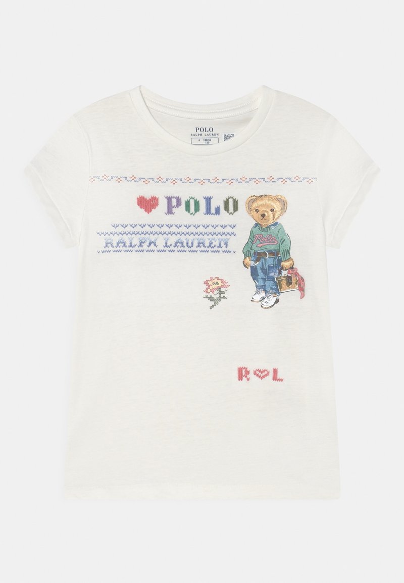 Biała dziecięca koszulka z krótkim rękawem z haftowanym tekstem "Polo Ralph Lauren", małą kwiatką oraz niedźwiedziem w zielonym swetrze, dżinsach i tenisówkach.