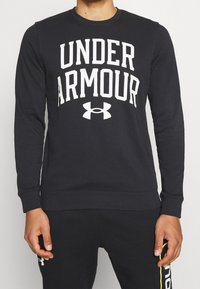 Felpa nera con grande testo bianco "UNDER ARMOUR" e logo sul davanti. Presenta un collo rotondo e polsini e orlo a coste.