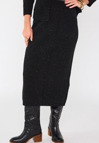 Jupe en maille noire avec des paillettes dorées, taille côtelée, associée à des bottes en cuir noires jusqu'au genou avec un talon épais marron.