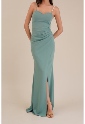 Kobieta ubrana w bezrękawnikową, zielonkawo-niebieską suknię wieczorową z marszczonym stanem, rozcięciem na udzie, perełkowymi ramiączkami i złotymi sandałami na wysokim obcasie na jednolitym tle.