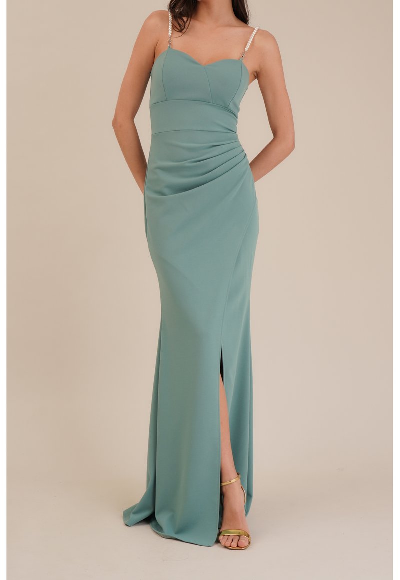 Femme portant une robe de soirée sans manches couleur sarcelle avec taille froncée, fente à la cuisse, bretelles en perles et sandales dorées à talons hauts sur fond uni.