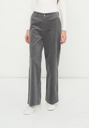 Personne portant un pantalon large en velours côtelé gris, un haut blanc à manches longues et des baskets blanches, debout devant un fond clair uni.