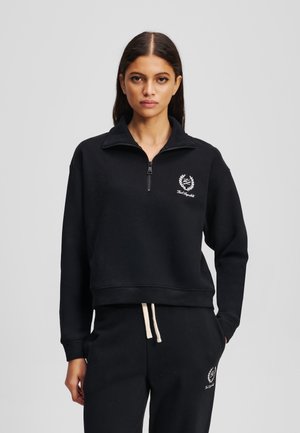 Sudadera de forro polar negra con media cremallera y cuello alto. Presenta un logo bordado blanco y puños elásticos. Combinada con pantalones deportivos negros.