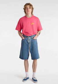 Rosa oversized t-shirt med gul logotyp, kombinerad med denimshorts med dragsko i midjan. Blå sneakers och vita strumpor kompletterar outfiten.