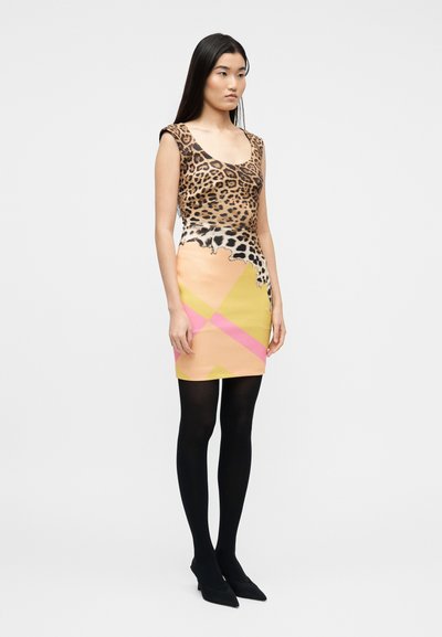 Γυναίκα που φοράει αμάνικο φόρεμα με τοπ λεοπάρ και φούστα με γεωμετρικά παστέλ σχέδια, μαύρο καλσόν και μαύρα μυτερά παπούτσια, στέκεται μπροστά σε λευκό φόντο.