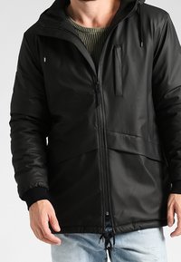 Man som bär en svart huva jacka med dragkedja över en olivgrön stickad tröja och ljusblå jeans.