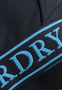 Gros plan d'une sangle en tissu bleu marine avec des lettres brodées bleu clair formant une partie d'un mot sur un fond noir texturé.