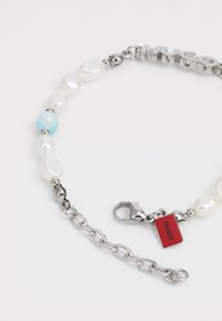 Brățară din lanț argintiu, decorată cu perle albe neregulate, o bilă aqua și un eticheta roșie cu "Hugo". Închizătoarea și linkurile oferă dimensiuni ajustabile.