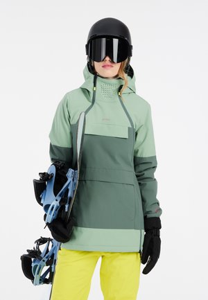 Giacca da snowboard impermeabile verde e grigia con cappuccio e tasca frontale; abbinata a pantaloni gialli e guanti neri; snowboard e attacchi in mano.