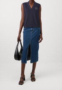 Námořnicky modrý top bez rukávů s výstřihem do V a logem, doplněný o vysoký denimový sukně s předním rozparkem, černou kabelkou a sandály na blokovém podpatku.