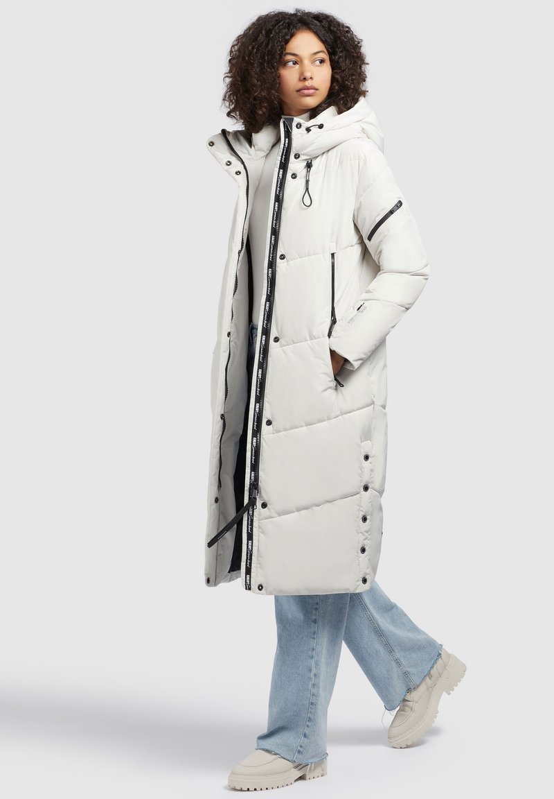Weiße lange Daunenjacke mit Kapuze, ausgestattet mit schwarzen Reißverschlüssen und Knöpfen, gestepptem Design und taillierter Passform. Kombiniert mit hellblauen Jeans und beigen Schuhen.