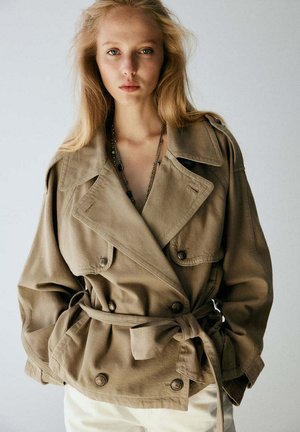 Joven con cabello largo y rubio que lleva una chaqueta beige de doble botonadura y grande atada a la cintura y pantalones de color claro sobre un fondo liso.