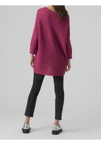 Femme debout de dos portant un long pull en tricot magenta, un jean noir slim et des chaussures à plateforme argentées.