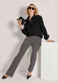 Frau in schwarzem Hemd und gemusterten Hosen, die an einem weißen Podest lehnt, eine schwarze Clutch hält, Sonnenbrille trägt und schwarze High Heels anhat.