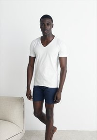 Un jeune homme debout pieds nus à l'intérieur, portant un t-shirt blanc à col en V et un short bleu marine, à côté d'un canapé beige contre un mur blanc.