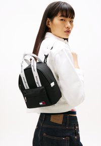 Μαύρο σακίδιο από νάιλον με στρογγυλεμένο σχήμα, λευκές λουρίδες με σήμα, μπροστινή τσέπη με φερμουάρ και πολύχρωμο πατς με το λογότυπο. Φοριέται στον ώμο.
