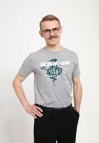 Šedé bavlněné tričko s grafickým potiskem "WORKER WEAR" v bílé a tyrkysové barvě, ve spojení s černými kalhotami a černým opaskem.
