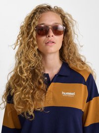 Vrouw met lang krullend blond haar, grote bruine zonnebril en een marineblauw en mosterd gestreept poloshirt met het logo "hummel sportswear".
