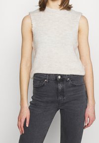 Světle béžový svetr bez rukávů s žebrovanou texturou a vysokým výstřihem. Spárováno se slim-fit tmavě šedými džíny.