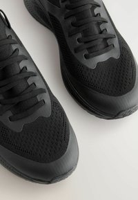 Baskets de sport noires avec une tige en maille texturée, des détails synthétiques et des lacets plats. Les semelles présentent un design de grip à motifs.